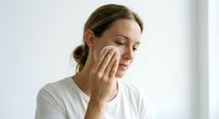 Young Caucasian woman gently applying skincare product with cotton pad on face in bright bathroom. Natural beauty routine and facial care concept.