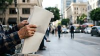 Mature man with brown skin holding blank newspaper on urban street with pedestrians and buildings in background, suitable for news, media, and advertising concepts.