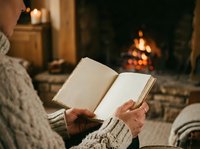 Close-up of person in cozy sweater reading open book by fireplace with warm ambient lighting and blurred flames in background.