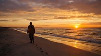 Male surfer in wetsuit walking along sandy beach at golden hour sunset with ocean waves and dramatic sky, suitable for travel and lifestyle content.