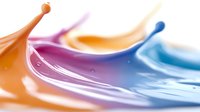 Colorful liquid paint drops colliding and splashing in dynamic motion creating abstract waves and curves with orange, purple and blue hues on white background.