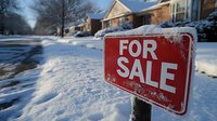 Red for sale sign covered in snow against suburban winter neighborhood background with snow covered yards and houses on residential street during cold season.