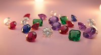 Colorful loose gemstones scattered on a pink surface, featuring rubies, emeralds, sapphires, amethysts, and diamonds sparkling in soft light.