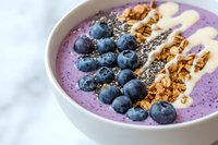 Purple smoothie bowl topped with fresh blueberries, banana slices, granola, and chia seeds on marble background, healthy breakfast concept for food blogs.