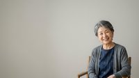Senior Asian woman with gray hair smiling at camera, wearing casual blue shirt and gray cardigan, seated against neutral background for portrait and lifestyle use.