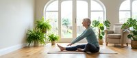 Senior woman with gray hair, Caucasian ethnicity, performing seated forward bend yoga pose on mat in bright living room with potted plants and large windows, wellness and home fitness.