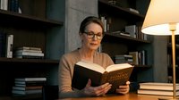 Mature Caucasian woman with glasses reading a book at a desk in a cozy library, warm lamp light, bookshelves in background, intellectual atmosphere.