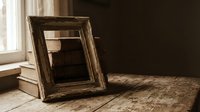 Vintage wooden picture frame with dark patina resting on rustic wooden surface near window, creating warm natural lighting for home decor and interior design.