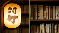 Traditional Japanese paper lantern glowing warmly beside wooden bookshelf filled with vintage Asian books and literature, creating atmospheric oriental ambiance.