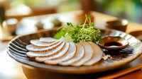 Sliced tender chicken breast garnished with fresh microgreens on black ceramic plate with soy sauce, served with wooden chopsticks in restaurant setting.