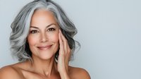 Mature woman with silver gray hair smiling confidently against light background, showcasing natural aging beauty and self-care for skincare advertising.
