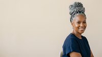 Mature African American woman with gray twisted hair styled in updo, wearing navy blue t-shirt, smiling at camera against neutral beige background, studio portrait for lifestyle and diversity content.