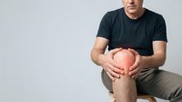 Young Caucasian man in dark t-shirt sitting and holding knee in pain, showing joint discomfort and injury on neutral background for medical concepts.