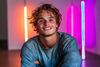 Young Caucasian man with curly hair smiling genuinely against colorful neon lights background. Wearing casual blue t-shirt in modern urban setting.