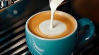 Barista pouring steamed milk into turquoise coffee cup creating heart latte art pattern, close-up view showing creamy foam texture and rich espresso blend.