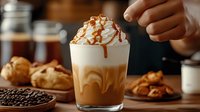 Layered caramel coffee drink topped with whipped cream and drizzle on rustic wooden table with coffee beans, pastries and nuts in warm lighting.