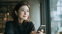 Young Asian woman using smartphone in modern cafe, looking thoughtful while browsing social media or texting, natural lighting from window creates warm atmosphere.