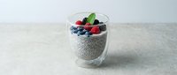 Chia seed pudding topped with fresh blueberries, raspberries, and mint leaf in glass cup on gray surface for healthy breakfast and wellness content.