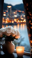 White roses in vase and glowing candle on windowsill against evening cityscape with bokeh lights reflecting on water, floral curtain detail creates cozy atmosphere.