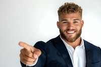 Confident young Caucasian businessman with beard and modern hairstyle pointing forward while smiling. Navy blazer and white shirt against light background.