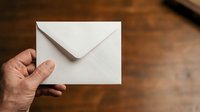Hand holding white envelope against wooden background, suitable for mail, correspondence, business communication, and postal service concepts.