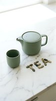 Sage green ceramic teapot and cup on marble surface with word tea spelled in small stones, minimalist zen style.