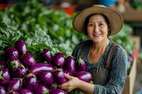 Mature woman farmer wearing straw hat and apron holding fresh purple eggplants at farmers market or garden stand with green foliage background.
