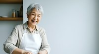 Senior Asian woman with white hair, wearing white apron and beige cardigan, smiling warmly in modern kitchen. Suitable for healthcare, wellness, cooking, and lifestyle content.