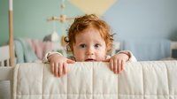Adorable toddler with curly red hair peeking over white crib rail in pastel nursery room. Child safety and bedroom furniture concept for parenting content.