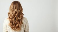 Young woman with long wavy blonde hair from behind wearing white knitted sweater against neutral background for hair care and beauty concepts.