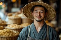 Young Asian farmer in traditional clothing and straw hat smiling at market stall with woven baskets of grain and agricultural products in background.