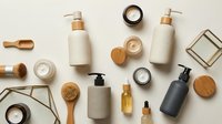 Flat lay arrangement of skincare and cosmetic products including pump bottles, jars, wooden brushes, and oil droppers on neutral background for beauty and wellness branding.