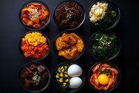 Korean side dishes in black bowls featuring kimchi, marinated meat, seasoned vegetables, boiled eggs and raw egg yolk on dark background. Traditional banchan set.