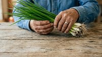 Mature man with weathered hands holding fresh green onions on wooden surface, wearing denim shirt, suitable for culinary, farming, and lifestyle content.