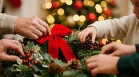 Hands crafting festive Christmas wreath with red ribbon bow and pine branches during holiday season preparation together.