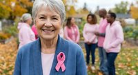 Senior Caucasian woman with gray hair wearing pink breast cancer awareness ribbon and blue cardigan smiling outdoors with support group in background.