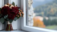 Elegant bridal bouquet with deep burgundy dahlias and peach roses rests on windowsill, natural light streaming through window creates romantic autumn atmosphere.