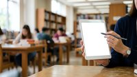 Woman in professional attire holding tablet with stylus in library or educational setting. Blurred colleagues working in background. Suitable for business, education, technology concepts.