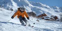 Young skier in bright orange jacket descending snowy mountain slope with alpine resort buildings and snow-covered peaks in background.