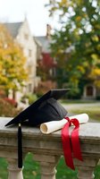 Graduation cap and diploma with red ribbon on stone ledge with blurred campus buildings and autumn trees in background for education celebration concepts.