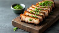 Sliced crispy pork belly with charred edges garnished with fresh chopped herbs on wooden serving board against dark background. Asian cuisine preparation.