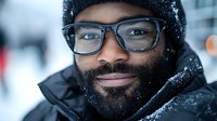 Young African American man with beard and glasses smiling warmly while wearing winter hat and coat during snowfall, expressing joy and confidence in cold weather.