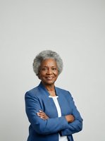 Senior African American woman with gray hair, wearing a blue blazer over white top, smiling confidently with arms crossed against neutral background. Corporate portrait for business, leadership, and professional use.