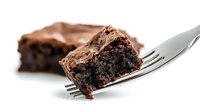 Moist chocolate brownie piece with rich fudgy frosting on silver fork against white background, close-up view showing dense texture and gooey center.