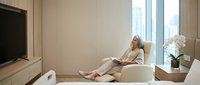 Mature Caucasian woman relaxing in modern beige armchair watching television in contemporary living room with city view through large windows.