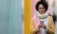 Young woman with curly hair and glasses wearing yellow cardigan and scarf checking smartphone against colorful urban wall background.