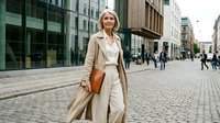 Confident mature businesswoman walking on urban street wearing elegant beige trench coat, carrying leather bag in modern city district for professional lifestyle.