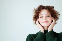 Young Caucasian woman with curly brown hair and natural makeup smiling warmly while holding face in hands, wearing green sweater against light background.