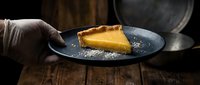 Hand in glove serving slice of golden custard tart on dark plate with powdered sugar dusting. Professional food presentation for bakery and dessert menus.