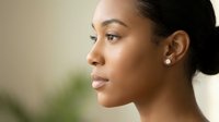 Young African American woman in profile with natural makeup and elegant pearl earrings against soft blurred background for beauty and skincare concepts.